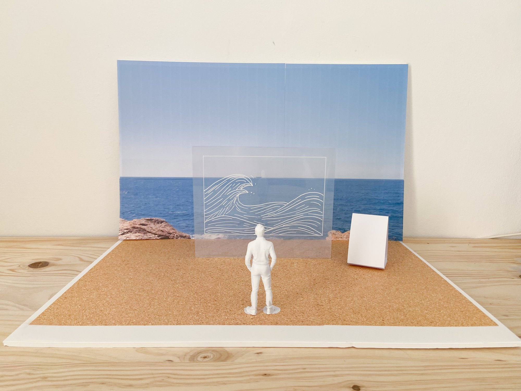 Maquette d'exposition : une feuille de plexiglas dessinée posée face à une photographie de la mer, figurine à l'échelle